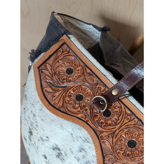 A tan leather cartwright hand-tooled bag patch with floral tooling and black stitches on hair-on hide background