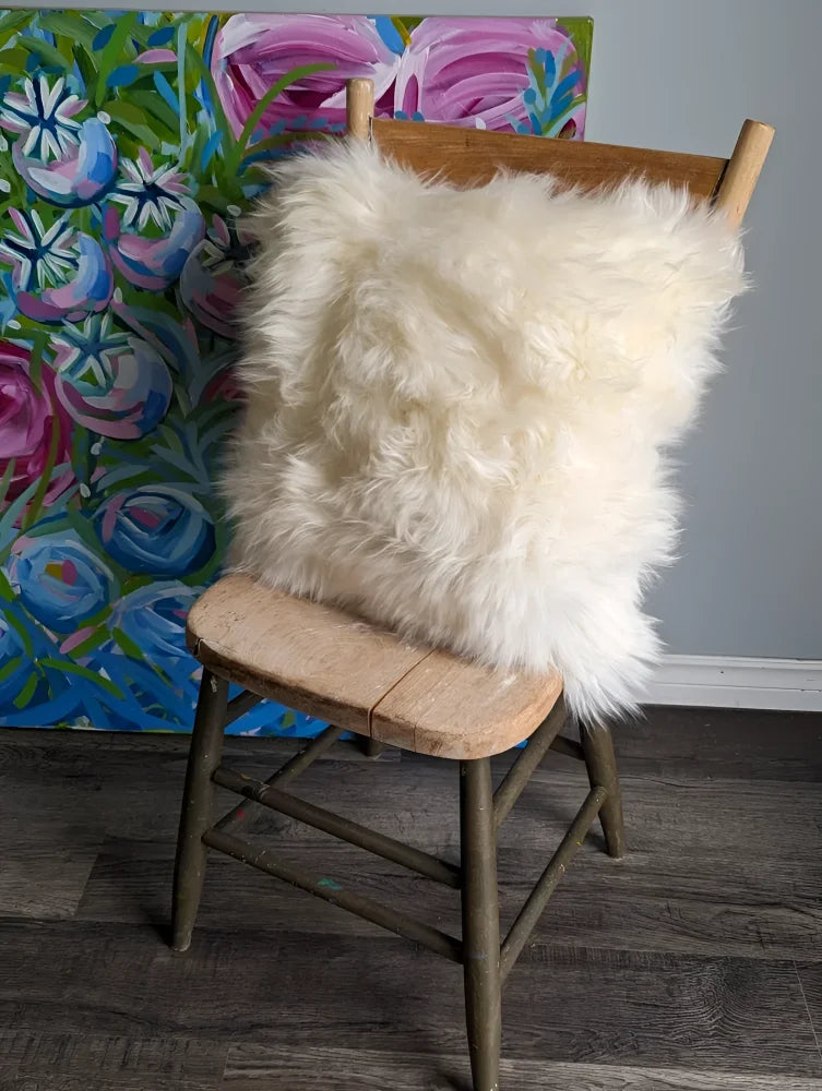 A plush off-white 16 x 16 merino sheepskin cushion cover with soft fluffy texture on a rustic wooden chair