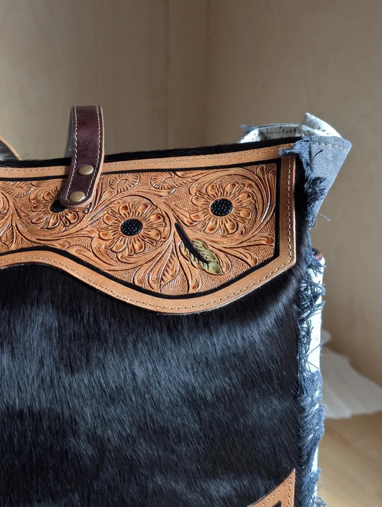 Black cowhide tote bag with tan leather floral tooling and green leaf accent featuring a zippered pocket and hair-on hide