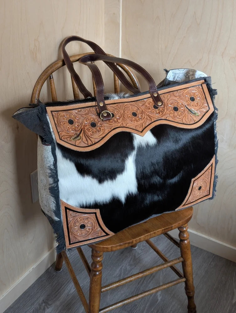 The sunflower weekender bag is a black and white hair-on hide tote with tan cartwright hand-tooled leather accents and a zippered pocket