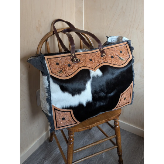The sunflower weekender bag is a black and white hair-on hide tote with tan cartwright hand-tooled leather accents and a zippered pocket