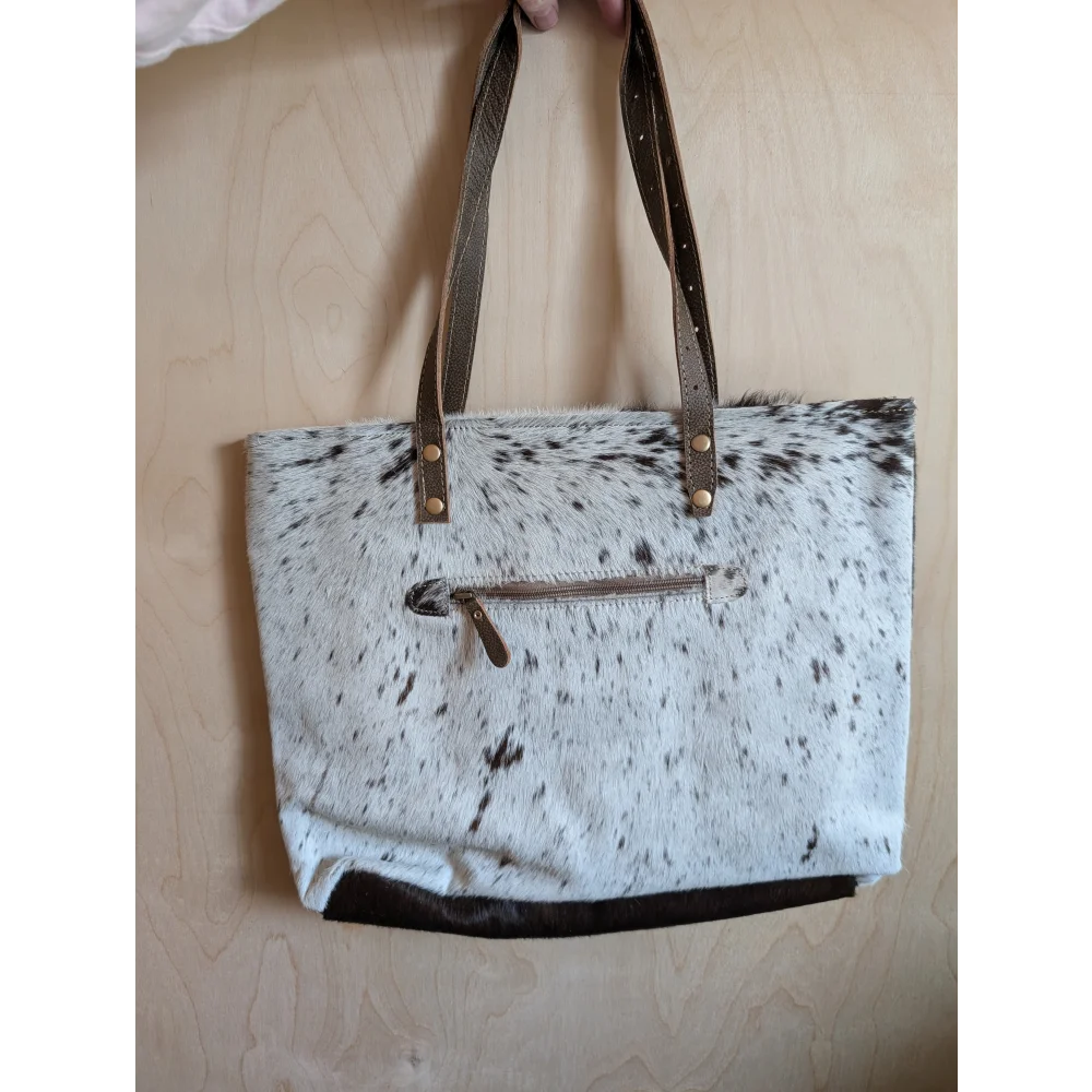 A white and black spotted real cowhide tote bag with brown leather straps and matching zipper pull from cowhide rugs collection