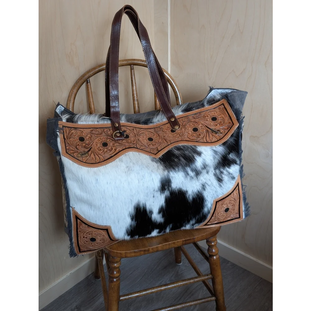 The sunflower weekender bag features a rustic cowhide hair-on hide design with tan leather embroidery and dark handles with zippered pocket
