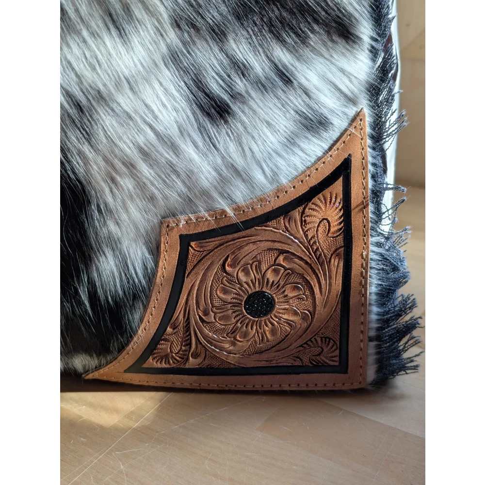 Brown leather cartwright hand-tooled bag with intricate floral design and black border on black-and-white hair-on hide background