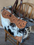 The sunflower weekender bag is a rustic hair-on hide cartwright hand-tooled bag with tan floral patterns and dark brown straps