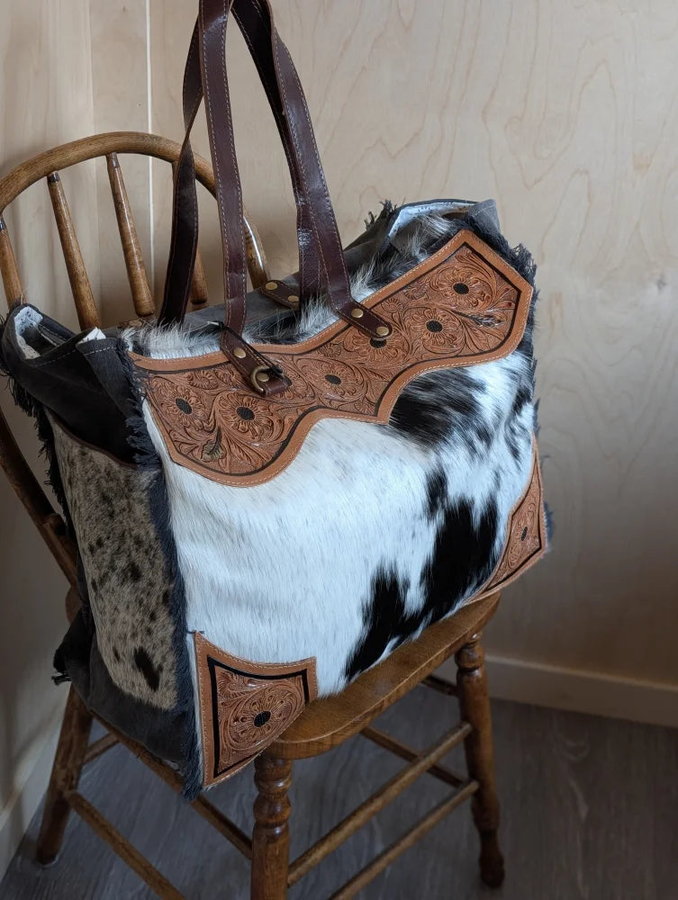 The sunflower weekender bag is a rustic hair-on hide tote with tan cartwright hand-tooled panels and dark brown handles with zippered pocket