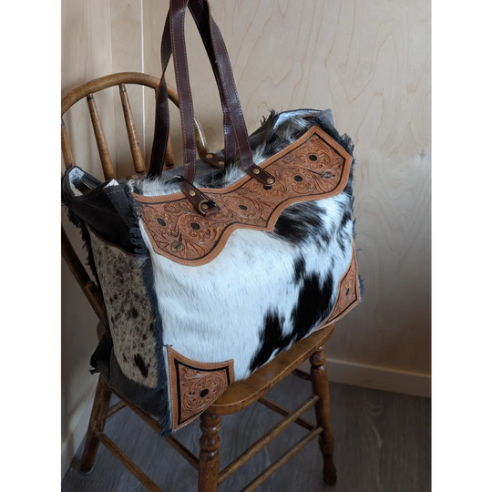 The sunflower weekender bag is a rustic hair-on hide tote with tan cartwright hand-tooled panels and dark brown handles with zippered pocket