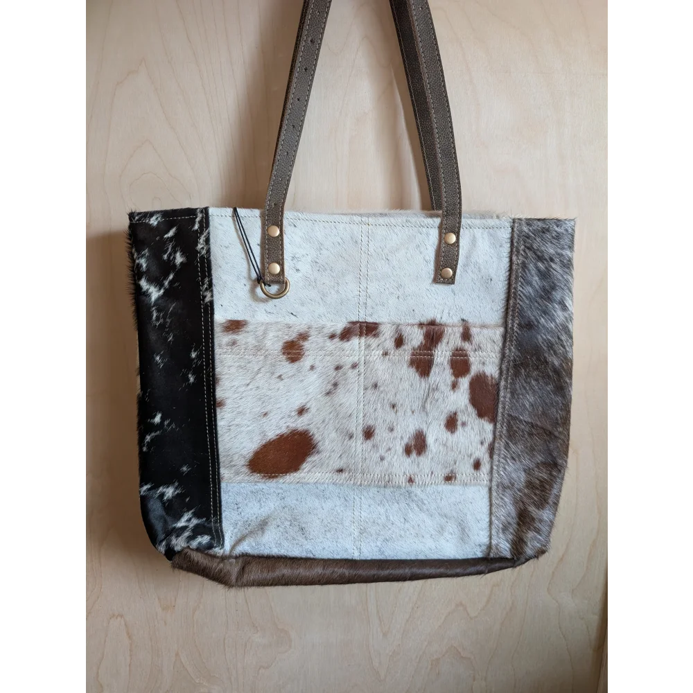 A stylish cowhide tote bag with patchwork white brown and black spots dark brown leather straps real cowhide design