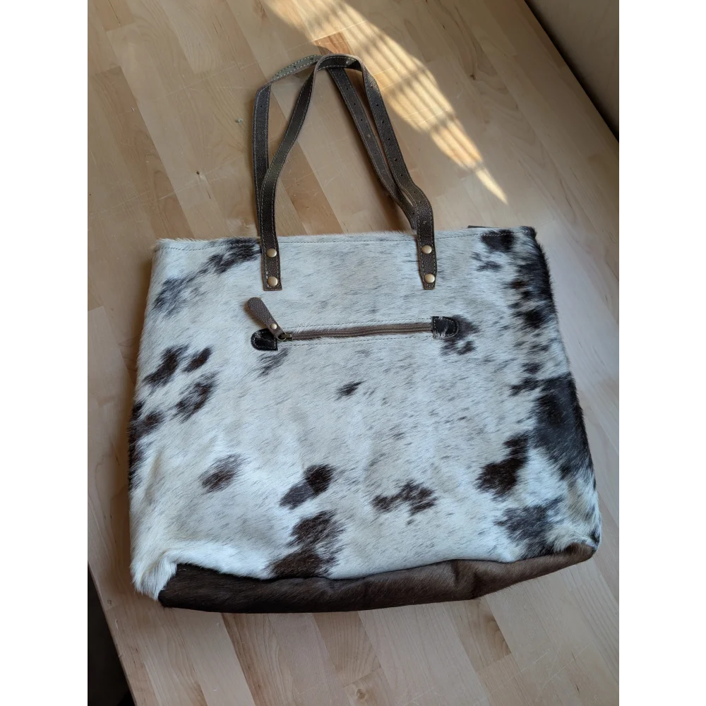 White and black cowhide tote bag with brown leather handles and zipper closure featuring real cowhide material