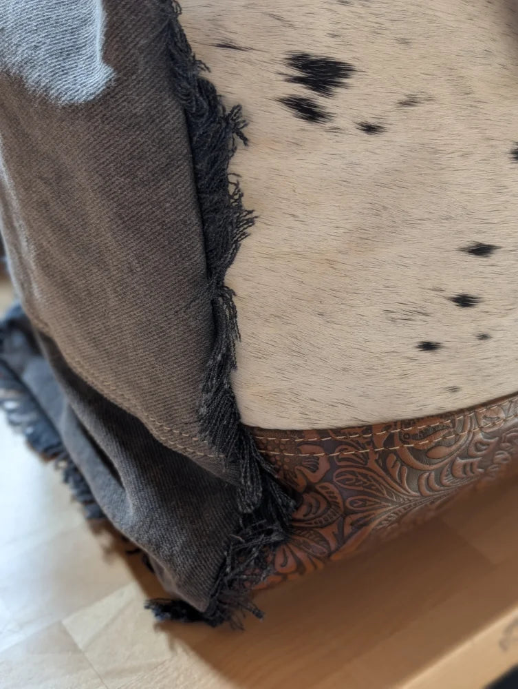 A rustic cowhide ottoman with cream base black spots frayed gray trim and leather accents inspired by matty affair canvas