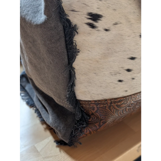 A rustic cowhide ottoman with cream base black spots frayed gray trim and leather accents inspired by matty affair canvas