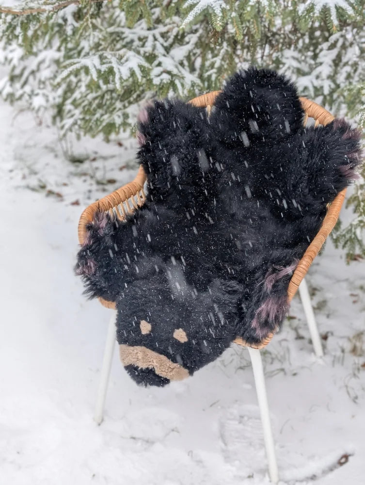 Adorable merino sheepskin black bear rug handmade plush with tan accents resting in a wicker chair outdoors in snowfall