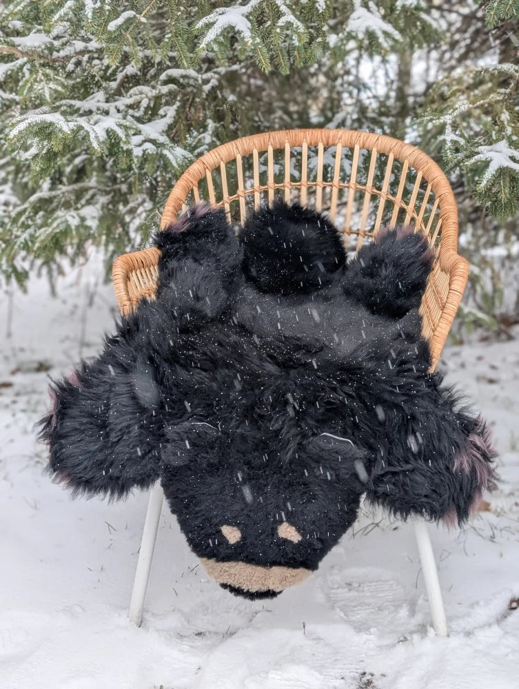 Adorable handmade merino sheepskin black bear rug with tan accents resting on wicker chair in snowy outdoors