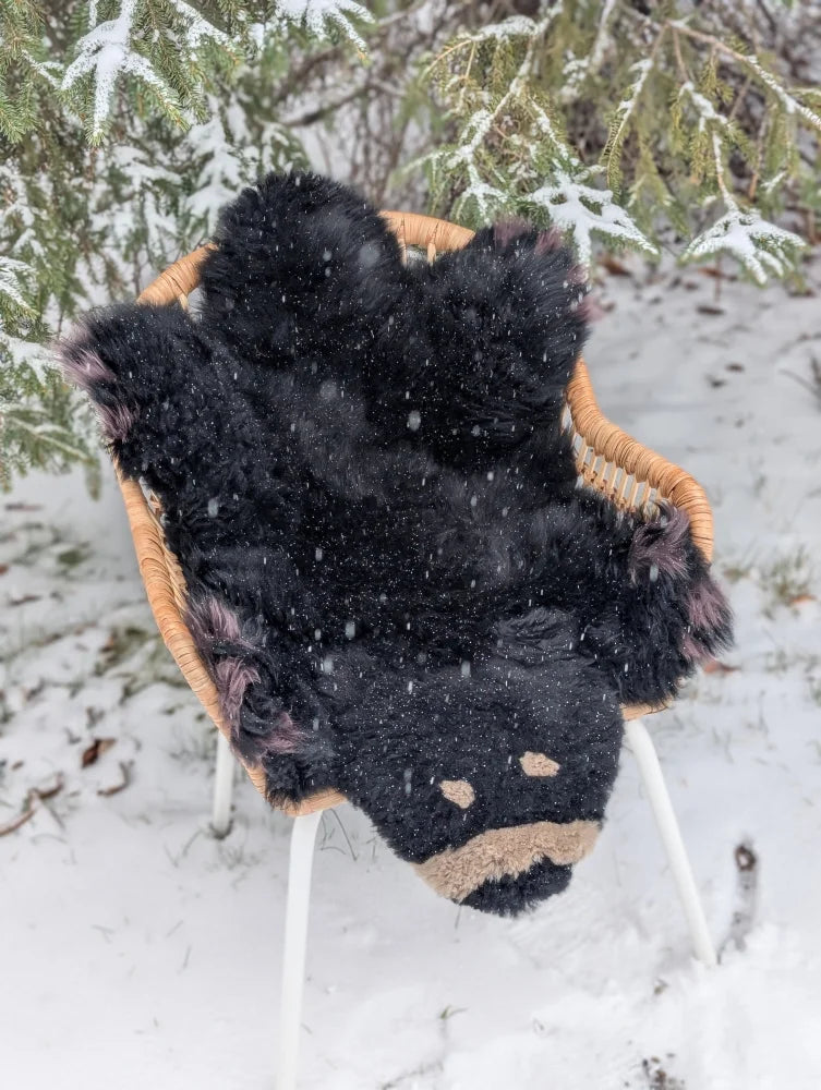 Adorable handmade merino sheepskin black bear rug with tan accents resting in a wicker chair outdoors