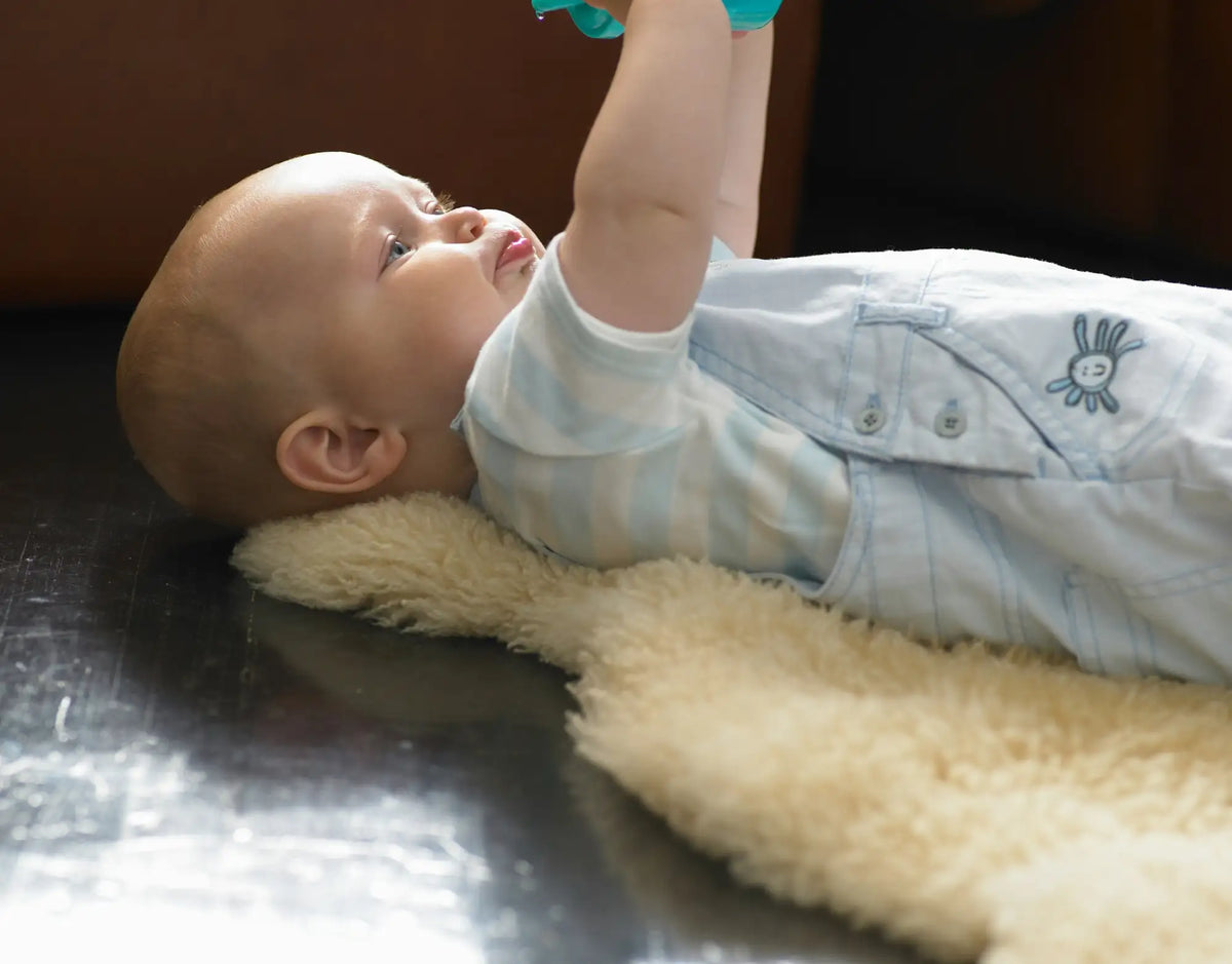 A baby wearing light blue and white striped overalls with a small embroidered sun design on the chest, lying on a soft beige sheepskin rug.