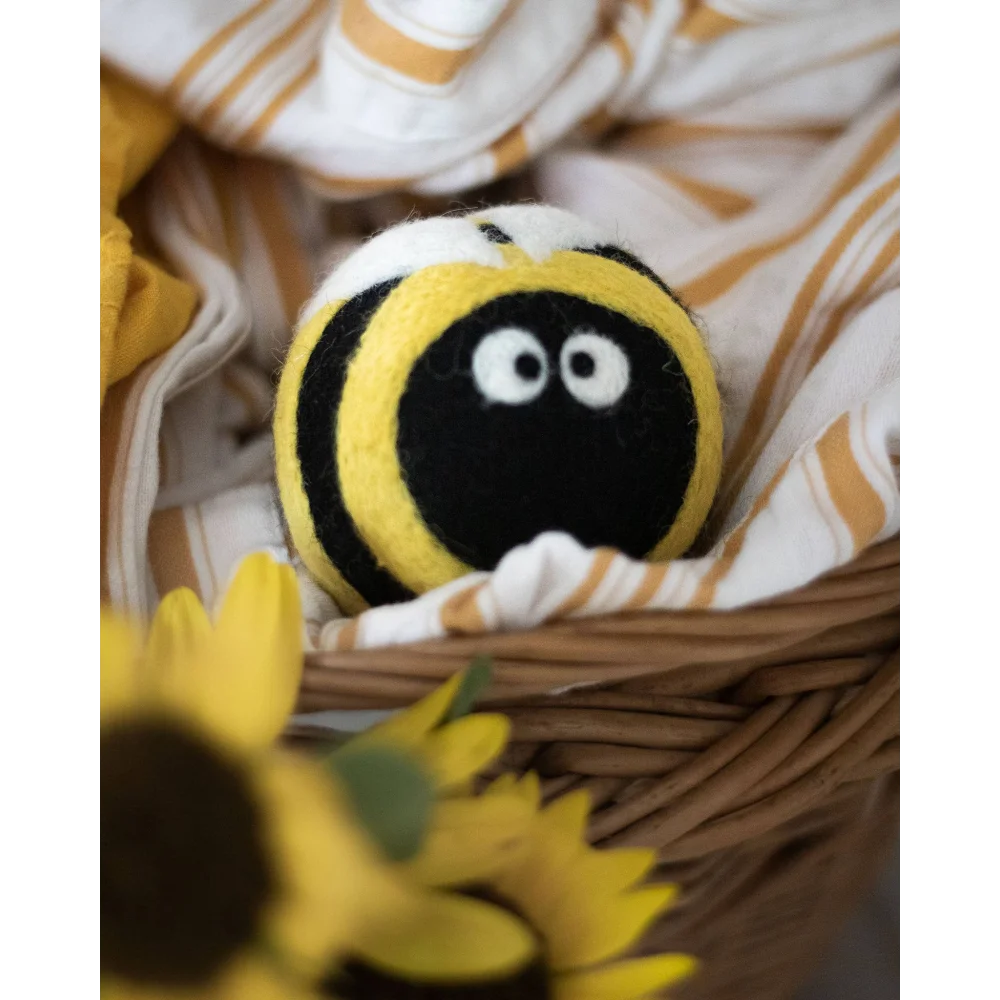 A plush felted bee dryer ball with black and yellow stripes and white eyes nestled in a woven basket eco dryer balls bee trio