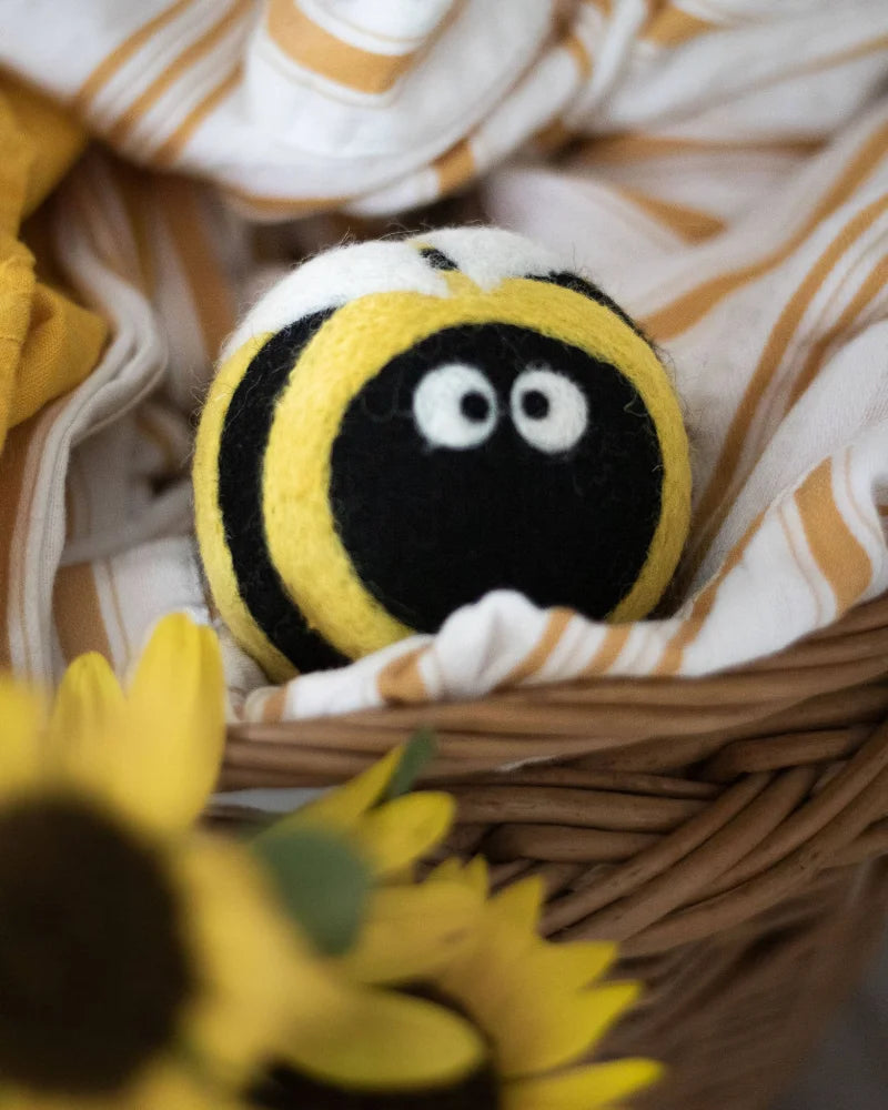 A plush felted bee dryer ball with black and yellow stripes and white eyes nestled in a woven basket eco dryer balls bee trio