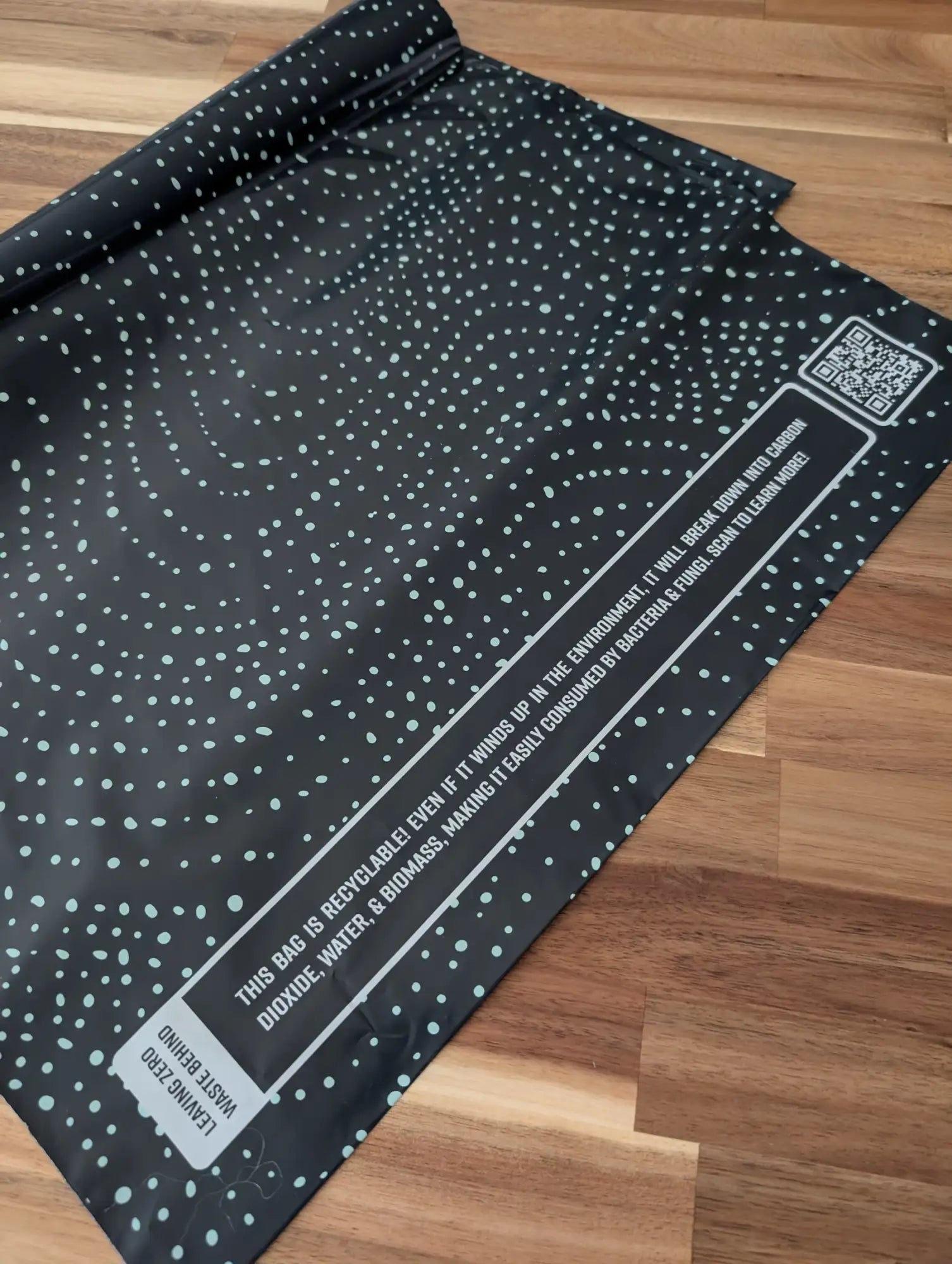 Black recycled mailer bag with a pattern of light teal dots and white text detailing its eco-friendly properties.