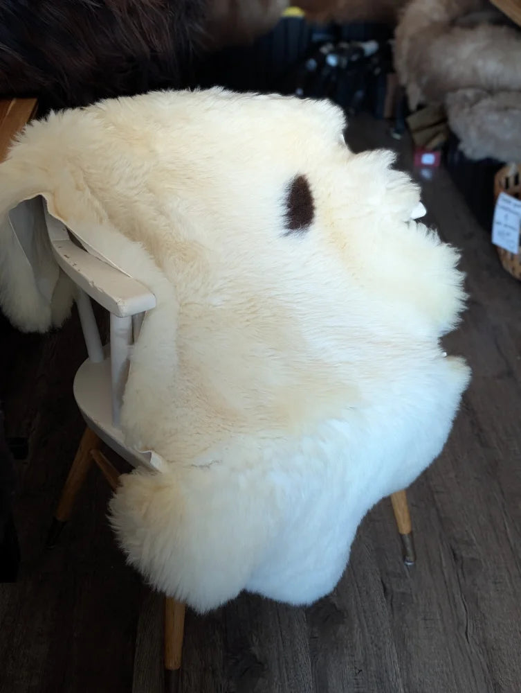 A plush off-white british isles sheepskin rug with a dark brown spot draped on a wooden chair for cozy decor