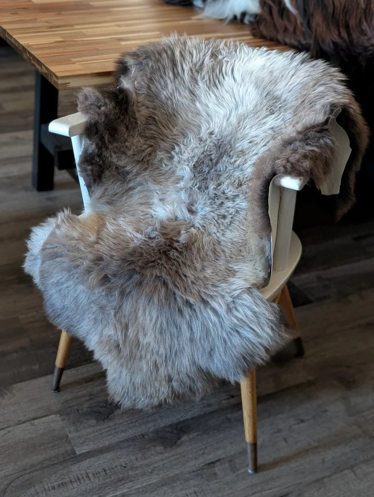 A plush british isles sheepskin rug in gray and brown draped over a wooden chair backrest for cozy decor