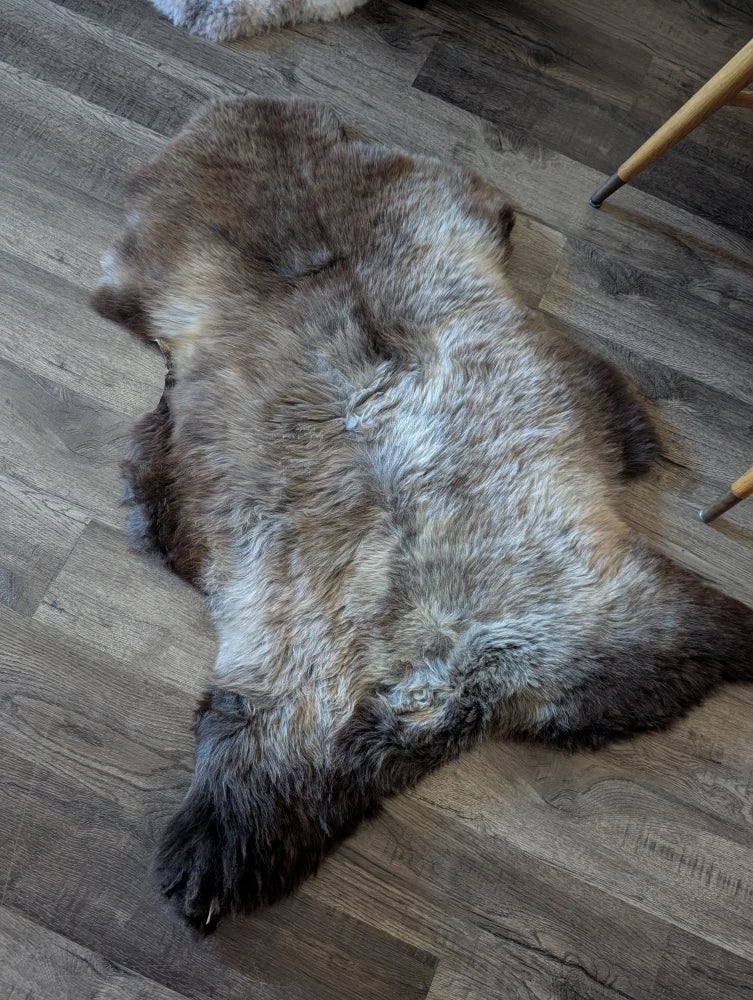A natural-toned british isles sheepskin rug with brown and gray hues lies flat on a wooden floor