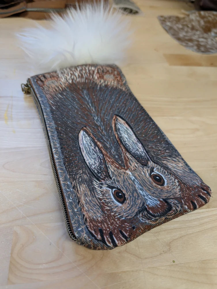 A hand-painted leather coin purse with two brown and gray rabbits and a fluffy white sheepskin tail pom pom