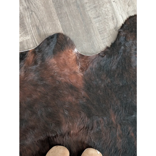 A large cowhide rug in dark brown and black with unique natural edges on a gray wood floor, perfect for beautiful rugs collections