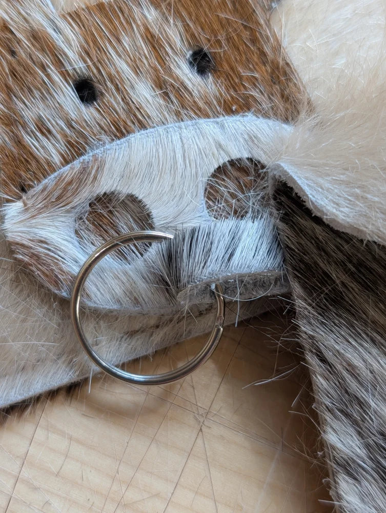 A silver metal keychain card holder with a simple ring design resting on brown and white cowhide surface