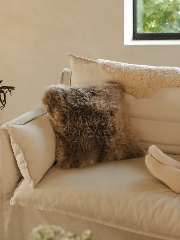 A plush shaggy brown merino sheepskin cushion with cotton backs resting on a cream sofa arriving october