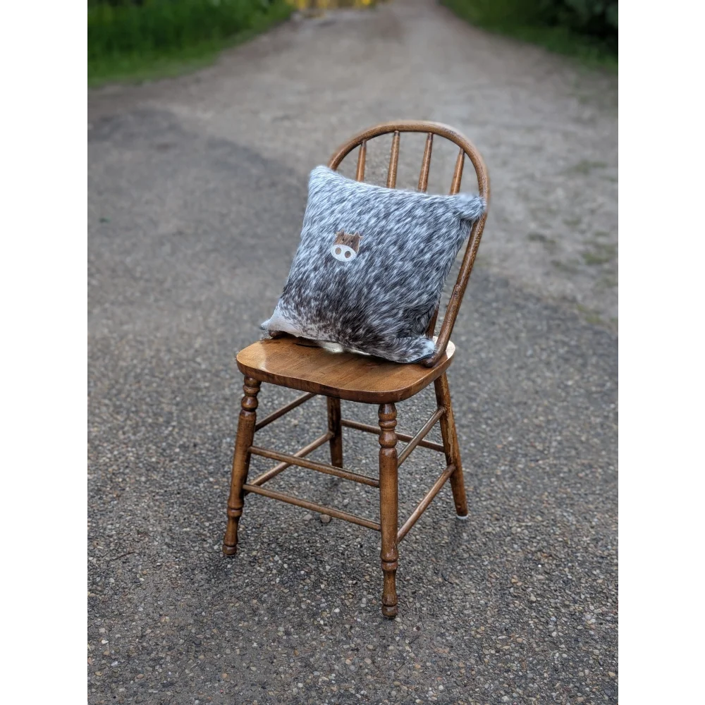 A cozy gray and white speckled mini moo double sided cowhide cushion with an embroidered animal face on a rustic wooden chair