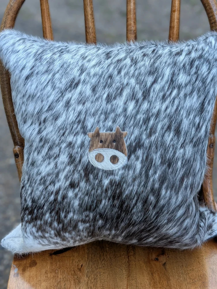 A cozy mini moo double sided cowhide cushion with gray and white speckled cowhide and a wooden cow face appliqué
