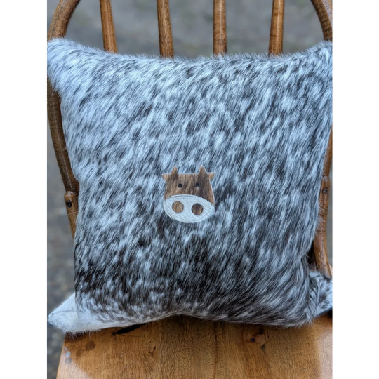 A cozy mini moo double sided cowhide cushion with gray and white speckled cowhide and a wooden cow face appliqué