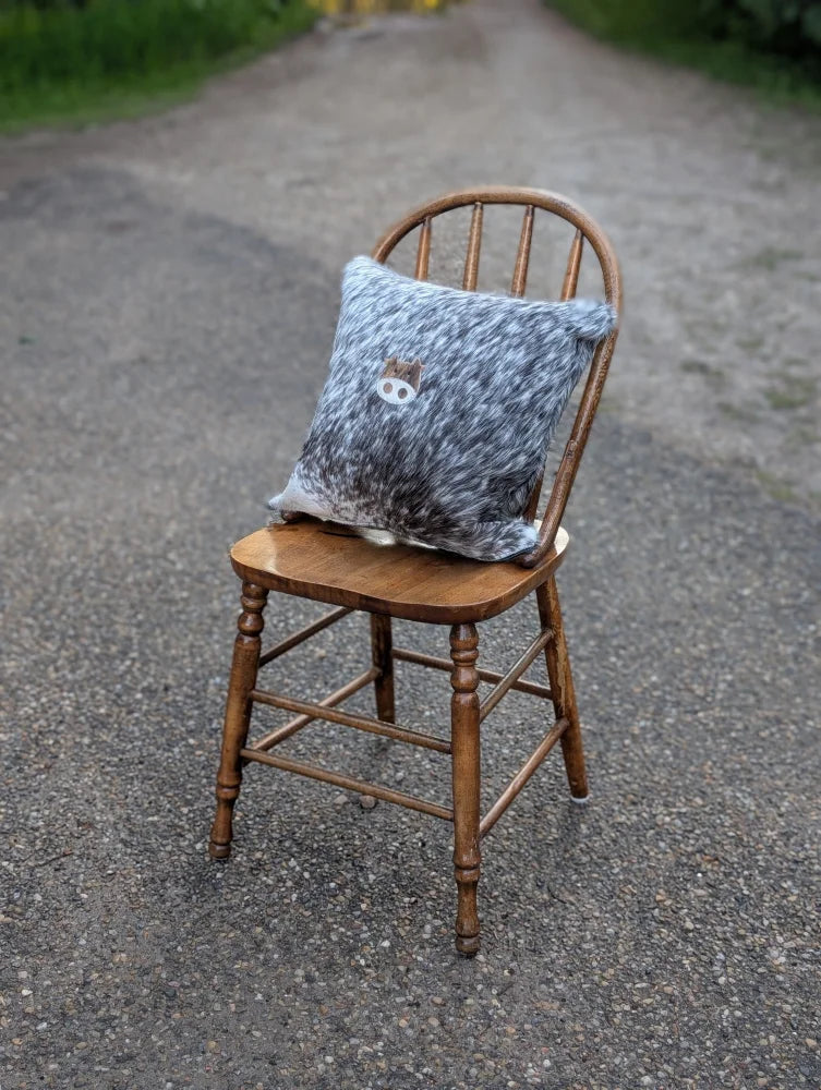A cozy gray and white speckled mini moo double sided cowhide cushion with an embroidered animal face on a rustic wooden chair
