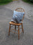A cozy gray and white speckled mini moo double sided cowhide cushion with an embroidered animal face on a rustic wooden chair