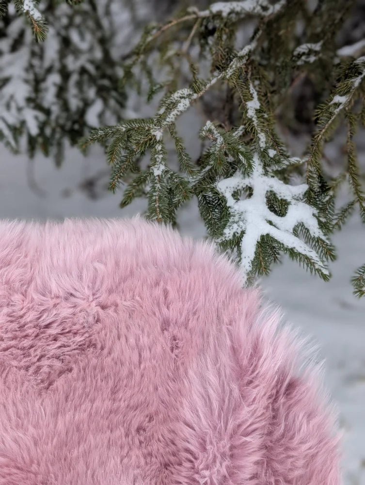 A soft fluffy pink merino sheepskin throw blanket rests against snow dusted evergreen branches meeting reach standards