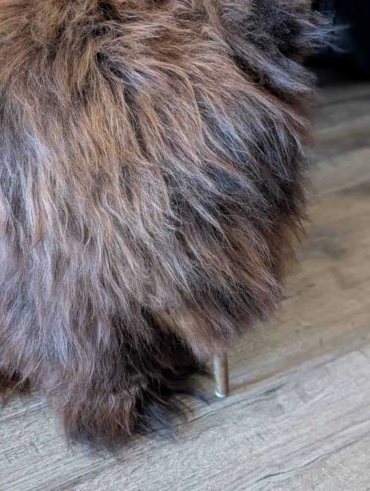 A plush stool with shaggy brown and gray icelandic sheepskin fur and a single metallic leg for cozy seating