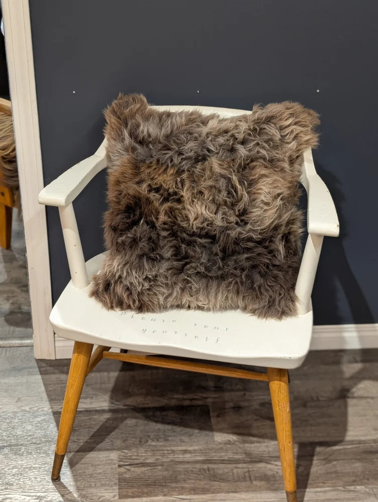A gorgeous british sheepskin cushion in plush brown rests on a white wooden chair with unbleached cotton canvas cover