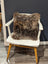 A gorgeous british sheepskin cushion in plush brown rests on a white wooden chair with unbleached cotton canvas cover