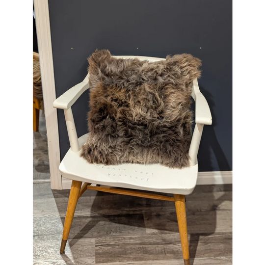 A gorgeous british sheepskin cushion in plush brown rests on a white wooden chair with unbleached cotton canvas cover