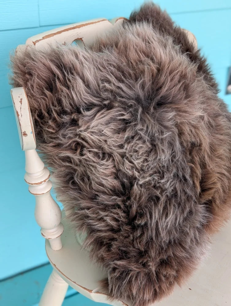 A plush shaggy brown british sheepskin cushion with soft curly texture on distressed white chair by angus oliver