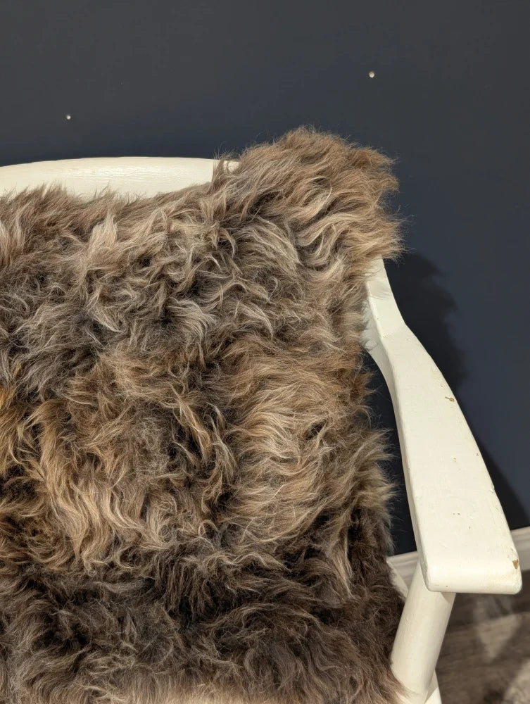 A plush curly brown british sheepskin cushion draped over a white wooden chair arm on unbleached cotton canvas