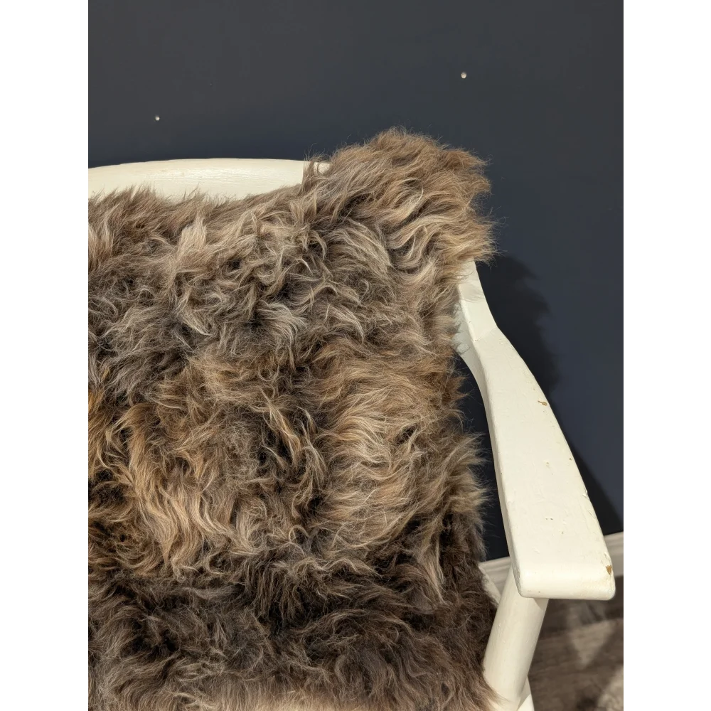 A plush curly brown british sheepskin cushion draped over a white wooden chair arm on unbleached cotton canvas