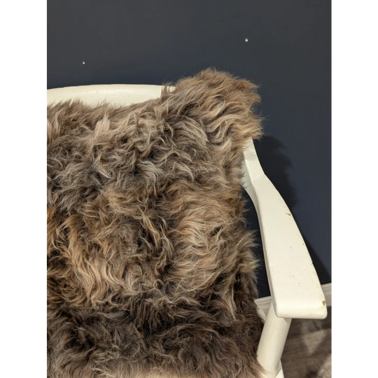 A plush curly brown british sheepskin cushion draped over a white wooden chair arm on unbleached cotton canvas