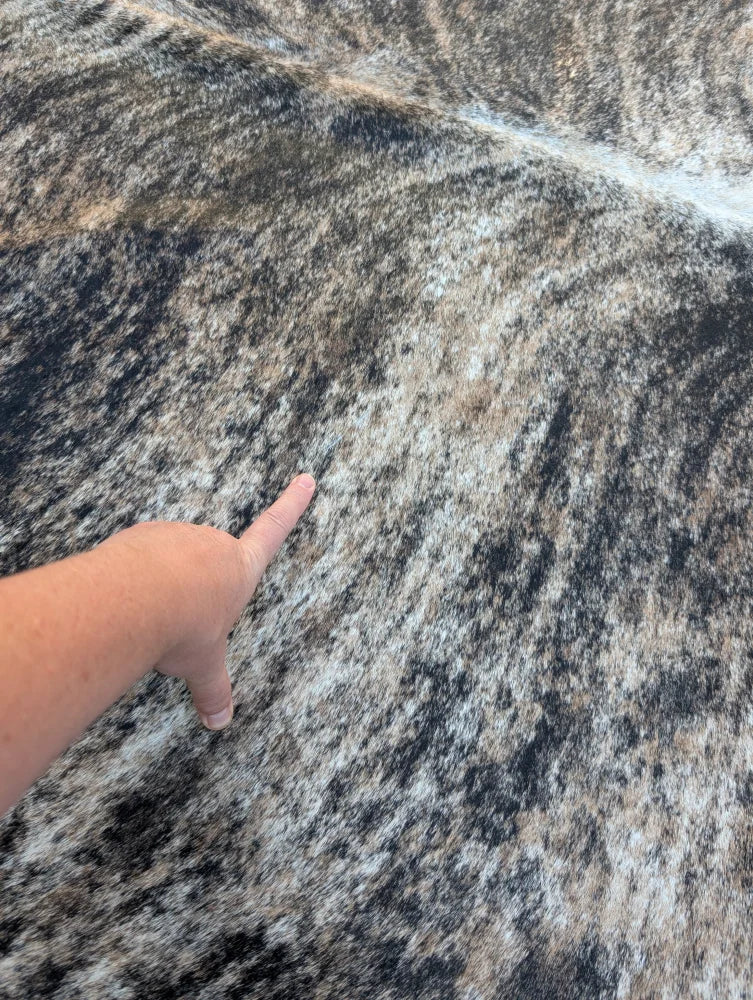 A hand points to a large cowhide rug with unique characteristics and rustic charm in black gray and beige tones