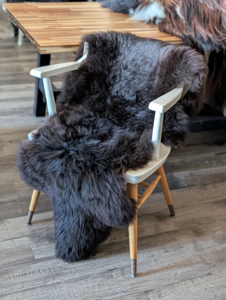 A plush dark gray british isles sheepskin rug draped over a light wooden chair for cozy comfort