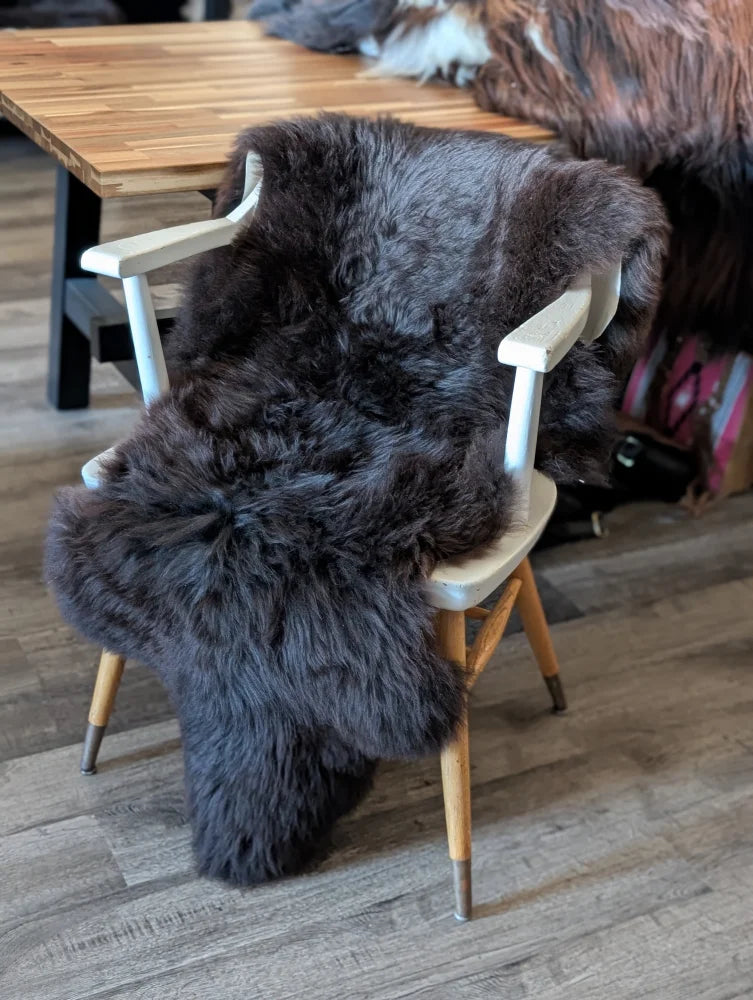 A plush dark gray british isles sheepskin rug draped over a white wooden chair with light wood legs