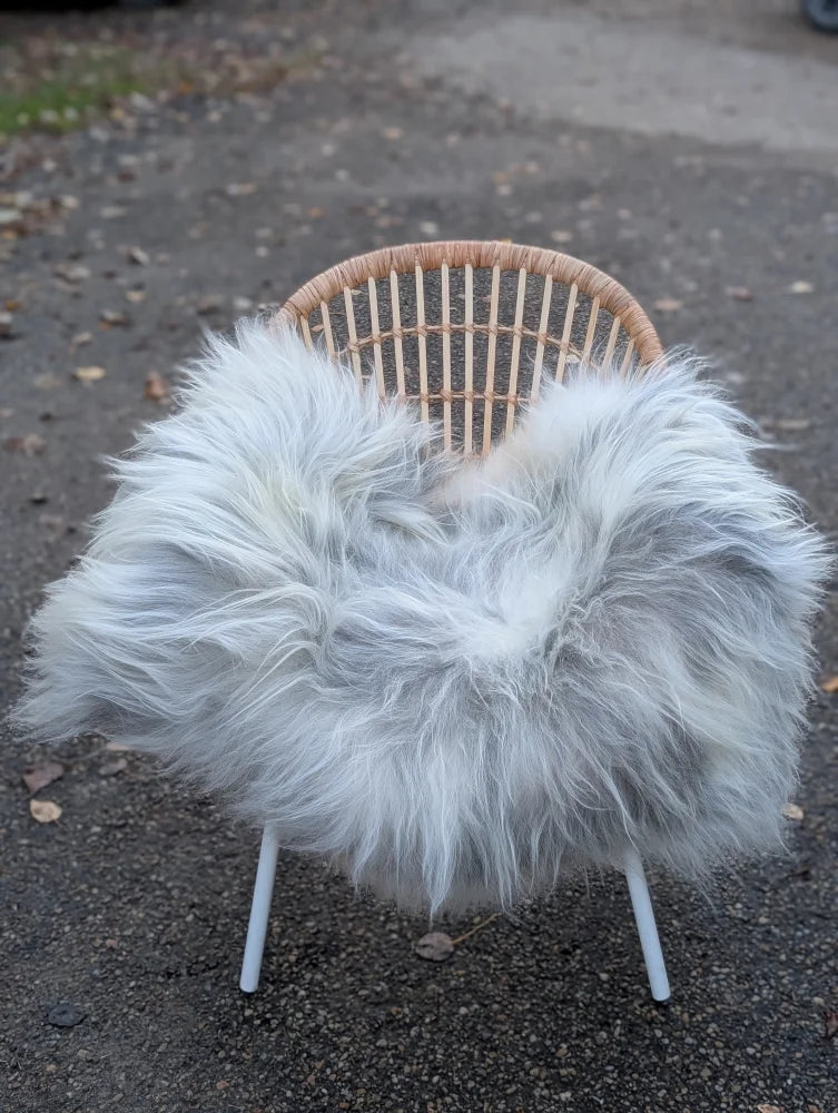A cozy chair with a woven wicker back and white legs draped in a plush grey icelandic sheepskin throw