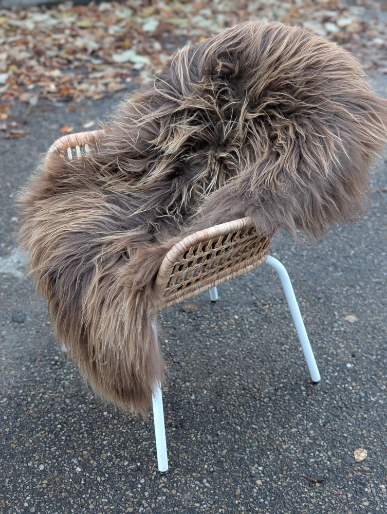 A cozy thick brown dark rusty icelandic sheepskin throw draped over a wicker chair with white metal legs