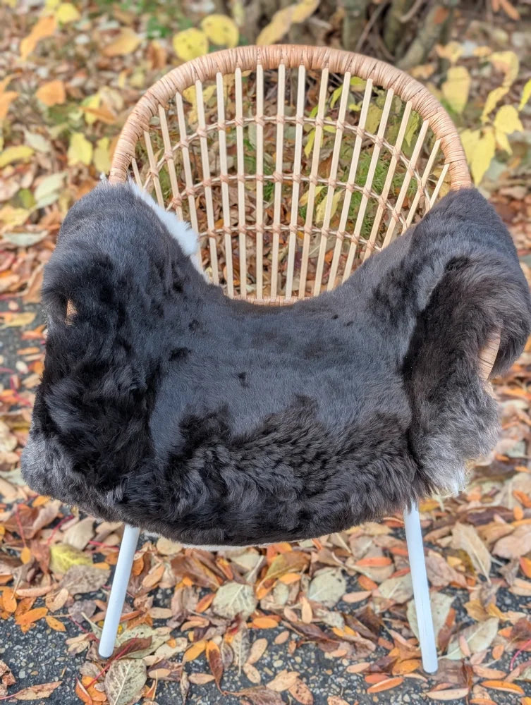 A plush dark gray wool icelandic sheepskin rug with dark edges draped over a woven wicker chair with white metal legs