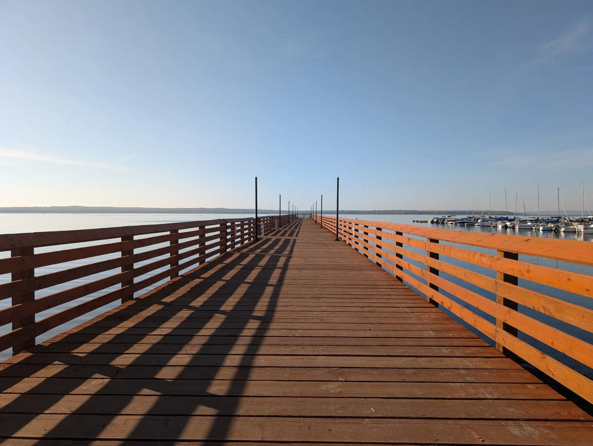 A long wooden pier with warm brown planks and matching railings stretches out over calm water under a clear blue sky.